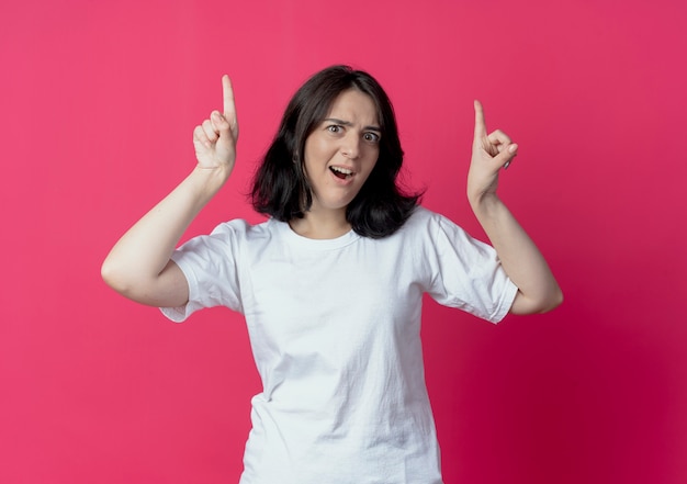 Impressed young pretty caucasian girl looking at camera and pointing up isolated on crimson background