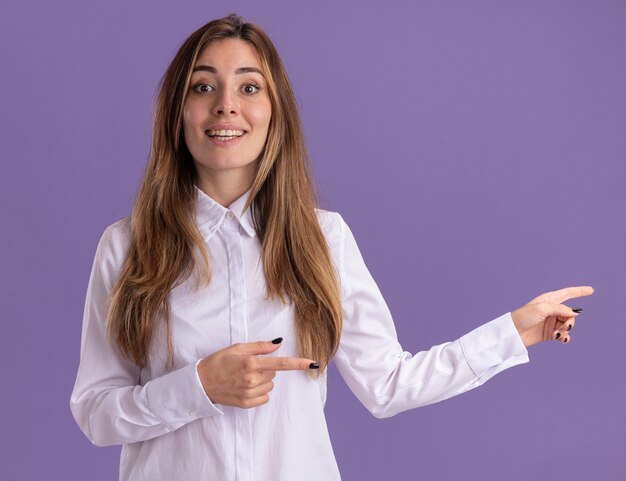 Впечатленная молодая симпатичная кавказская девушка указывает в сторону двумя руками на фиолетовом