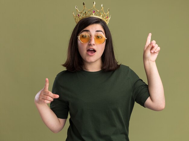 Впечатленная молодая симпатичная кавказская девушка в солнцезащитных очках с короной, указывающей вверх двумя руками на оливково-зеленом