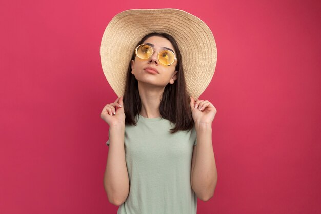 Impressed young pretty caucasian girl wearing beach hat and sunglasses grabbing hat looking up 