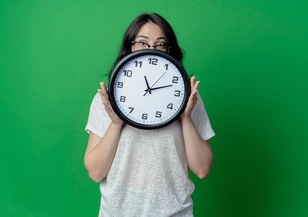 Впечатленная молодая симпатичная кавказская девушка в очках держит часы и смотрит в камеру сзади, изолированную на зеленом фоне с копией пространства
