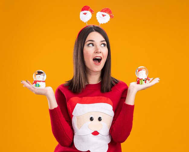 Impressed young pretty caucasian girl wearing santa claus headband and sweater holding snowman and santa claus figurines looking at side isolated on orange background