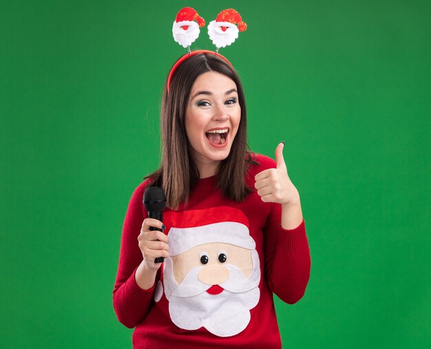 Impressed young pretty caucasian girl wearing santa claus sweater and headband holding microphone  showing thumb up isolated on green wall with copy space