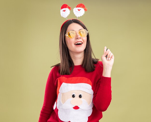 Impressed young pretty caucasian girl wearing santa claus sweater and headband with glasses looking and pointing up isolated on olive green background with copy space
