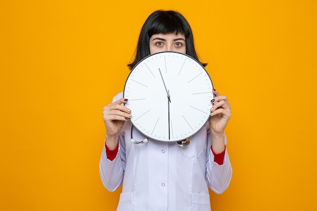 Free photo impressed young pretty caucasian woman in doctor uniform with stethoscope holding clock in front of her face