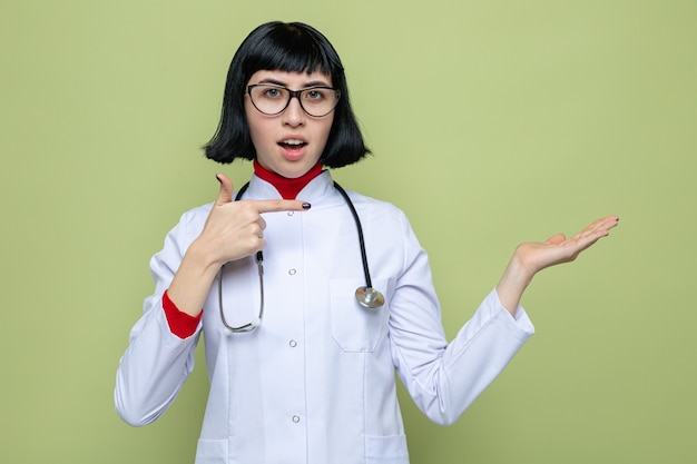 Impressionata giovane bella donna caucasica in uniforme da medico con uno stetoscopio che punta alla sua mano vuota