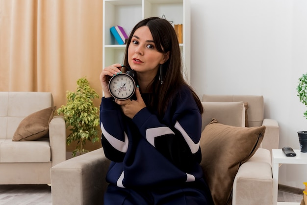 Impressed young pretty caucasian woman sitting on armchair in designed living room holding alarm clock looking