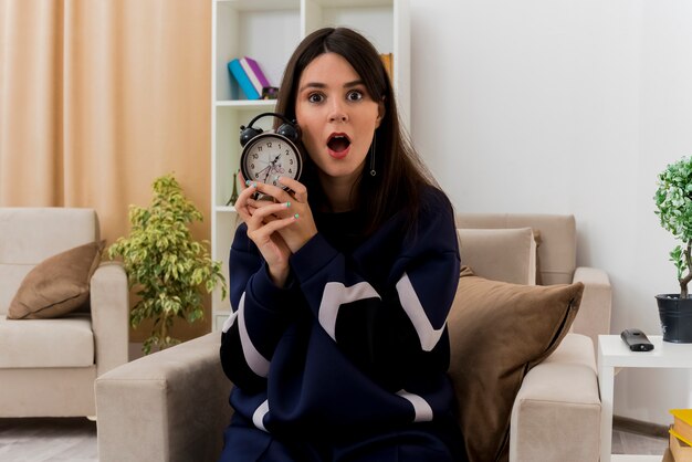 Impressed young pretty caucasian woman sitting on armchair in designed living room holding alarm clock looking