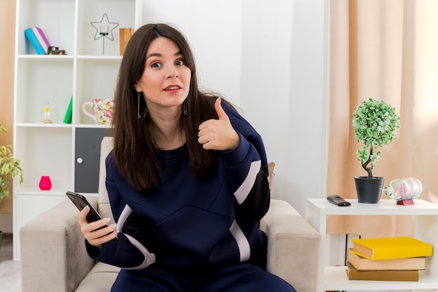 Impressed young pretty caucasian woman sitting on armchair in designed living room holding mobile phone looking and showing thumb up