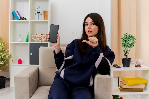 Impressed young pretty caucasian woman sitting on armchair in designed living room showing mobile phone and pointing at it