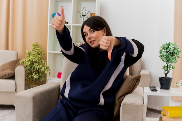 Impressed young pretty caucasian woman sitting on armchair in designed living room showing thumbs up and down