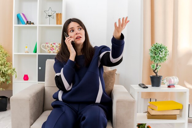 Impressed young pretty caucasian woman sitting on armchair in designed living room talking on phone looking at side and showing empty hand