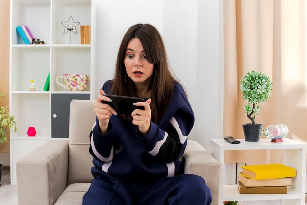 Impressed young pretty caucasian woman sitting on armchair in designed living room watching something on phone