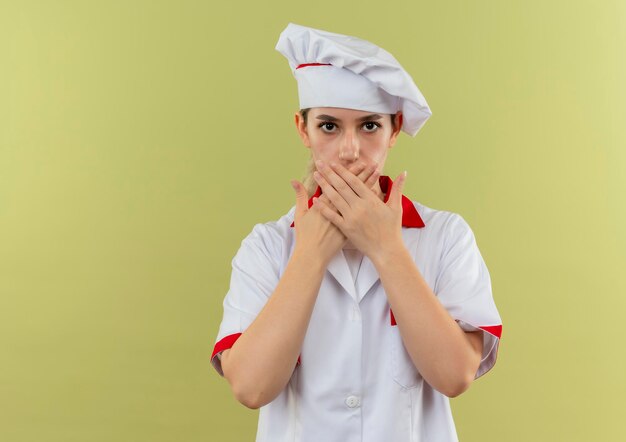 Impressed young pretty cook in chef uniform closing mouth with hands isolated on green wall with copy space