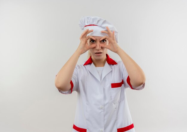 Impressed young pretty cook in chef uniform doing look gesture using hands as binoculars isolated on white wall