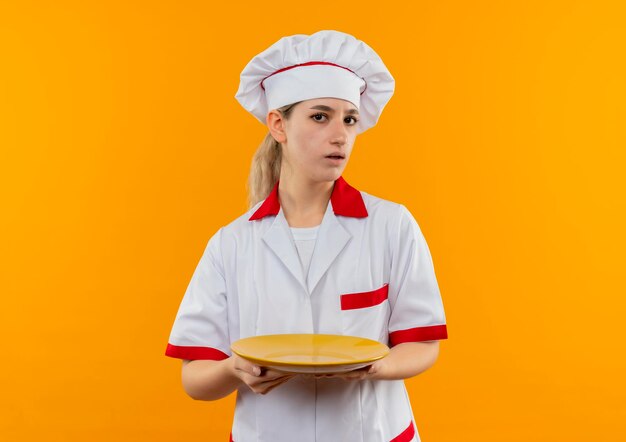 Impressed young pretty cook in chef uniform holding empty plate  isolated on orange wall