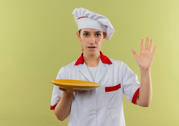 空の皿を持ち、緑の壁に手を上げてシェフの制服を着た若いかわいい料理人
