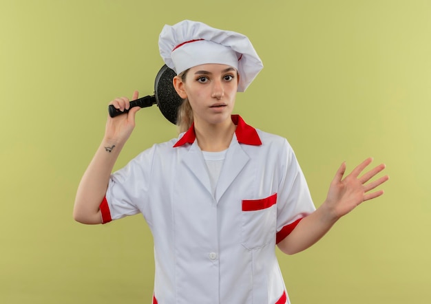 그녀의 머리 뒤에 프라이팬을 들고 녹색 벽에 고립 된 빈 손을 보여주는 요리사 유니폼에 감동 된 젊은 예쁜 요리사