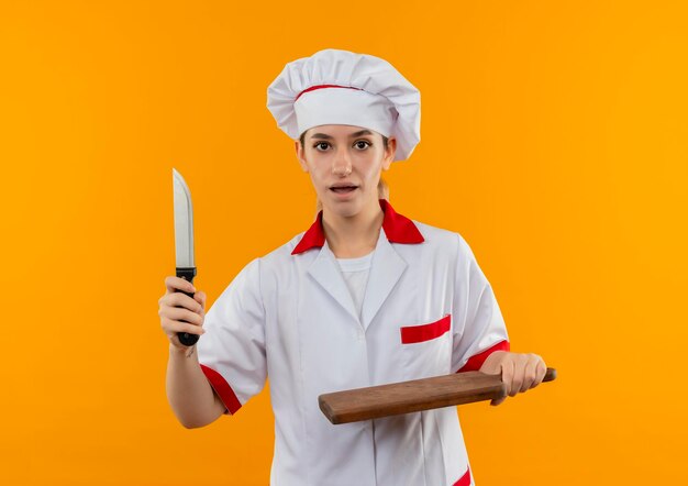 오렌지 벽에 고립 된 칼과 커팅 보드를 들고 요리사 유니폼에 감동 된 젊은 예쁜 요리사