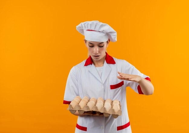 Foto gratuita impressionato giovane bella cuoca in uniforme da chef che tiene e guarda il cartone di uova tenendo la mano in aria isolata sulla parete arancione con spazio di copia