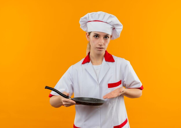 オレンジ色の壁に分離されたフライパンを保持し、指差すシェフの制服を着た若いかわいい料理人