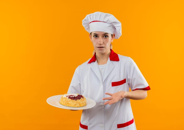 Impressed young pretty cook in chef uniform holding and pointing with hand at plate of cake isolated on orange wall