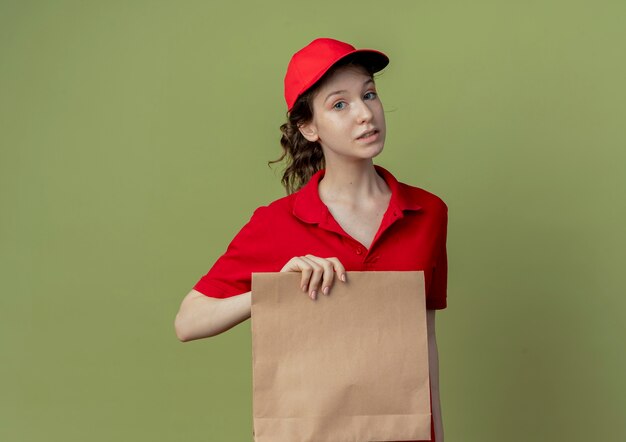 Впечатленная молодая симпатичная доставщица в красной форме и кепке, держащая бумажный пакет, смотрящую в камеру, изолированную на оливково-зеленом фоне с копией пространства