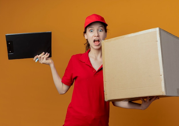 Impressed young pretty delivery girl in red uniform and cap holding pen and clipboard with carton box looking down isolated on orange background