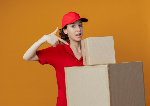 빨간 유니폼과 모자를 들고 오렌지 배경에 고립 된 판지 상자를 가리키는 감동 된 젊은 예쁜 배달 소녀