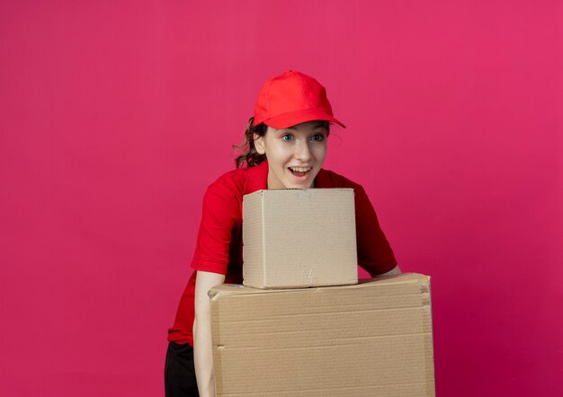 Impressed young pretty delivery girl in red uniform and cap looking straight holding carton boxes isolated on crimson background with copy space