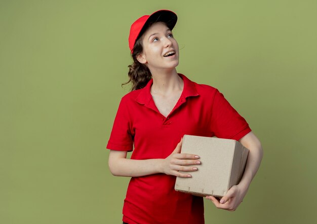 빨간 유니폼과 모자를 찾고 복사 공간이 올리브 녹색 배경에 고립 된 판지 상자를 들고 감동 젊은 예쁜 배달 소녀