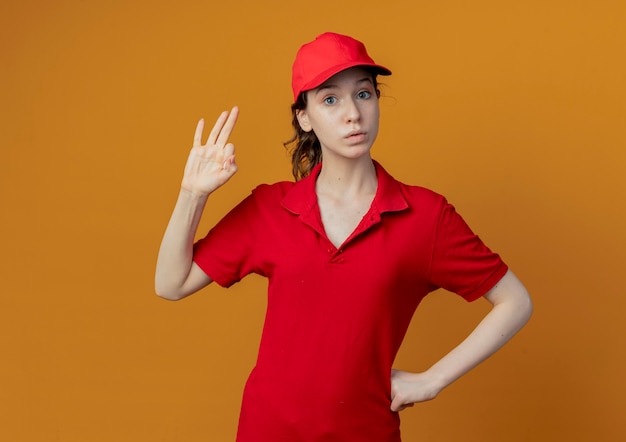 Impressed young pretty delivery girl in red uniform and cap putting hand on waist and doing ok sign