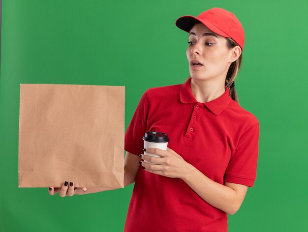 impressed young pretty delivery girl in uniform holds paper cup and looks at paper package on green