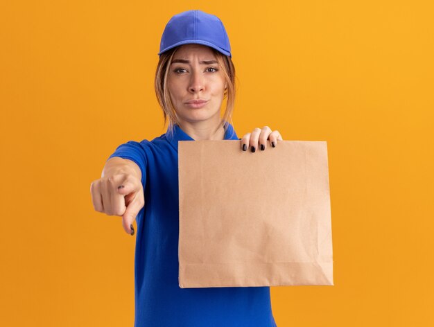 Impressed young pretty delivery girl in uniform holds paper package and points at camera on orange