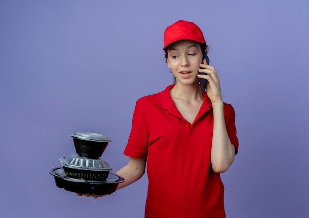 赤い​制服​と​帽子​を​身​に​着けて​食品​容器​を​保持し​、​見て​、​コピー​スペース​で​紫色​の​背景​に​分離された​電話​で​話している​印象的​な​若い​かわいい​配達​の​女の子