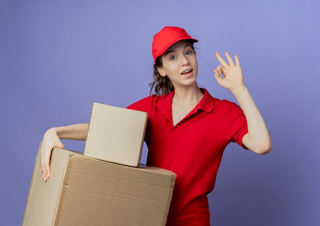 Impressed young pretty delivery girl wearing red uniform and cap holding carton boxes and doing ok sign isolated on purple background