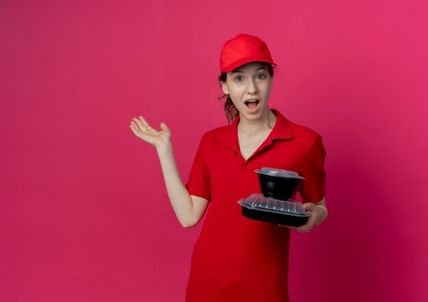 빨간 유니폼과 모자 복사 공간이 진홍색 배경에 고립 된 빈 손을 보여주는 음식 용기를 들고 감동 젊은 예쁜 배달 소녀