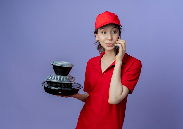 Impressed young pretty delivery girl wearing red uniform and cap looking at side holding food containers and talking on phone isolated on purple background with copy space