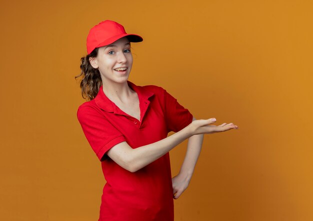 impressed young pretty delivery woman in red uniform and cap pointing with hand at side