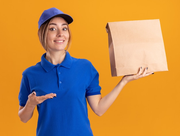 Impressed young pretty delivery woman in uniform holds and points at paper package with hand isolated on orange wall