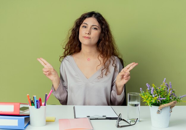 Impressed young pretty female office worker sitting at desk with office tools pointings at sides isolated on olive background