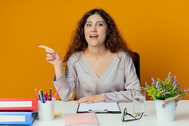 Impressed young pretty female office worker sitting at desk with office tools points at side isolated on orange wall with copy space