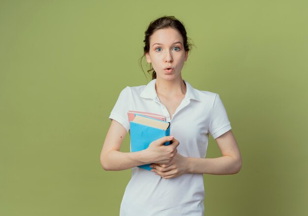 Impressed young pretty female student holding book and note pad isolated on olive green background with copy space