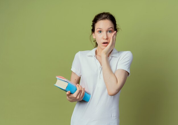 Impressed young pretty female student holding book and note pad putting hand on face isolated on olive green background with copy space