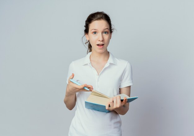 Впечатленная молодая симпатичная студентка держит открытую книгу и ручку, изолированные на белом фоне с копией пространства