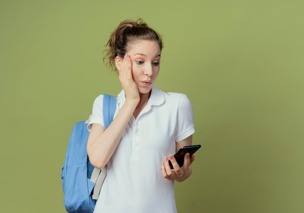 Impressed young pretty female student wearing back bag holding and looking at mobile phone and putting hand on face isolated on green background with copy space