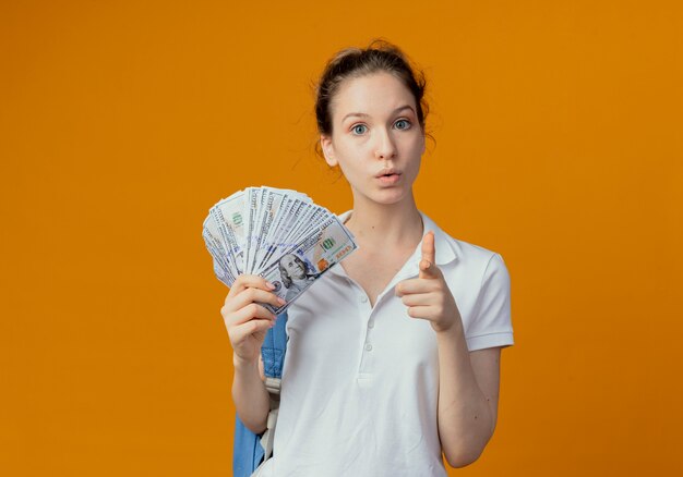 Impressed young pretty female student wearing back bag holding money and pointing at camera isolated on orange background with copy space