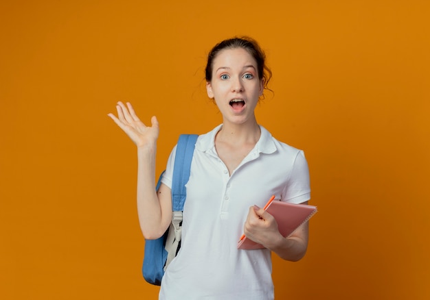 Impressed young pretty female student wearing back bag holding note pad and pen showing empty hand isolated on orange background with copy space