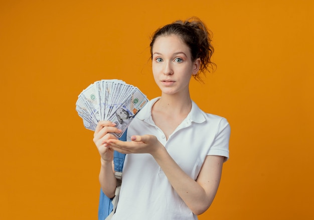 Впечатленная молодая симпатичная студентка в задней сумке, держащая и указывающая рукой на деньги, изолированные на оранжевом фоне с копией пространства