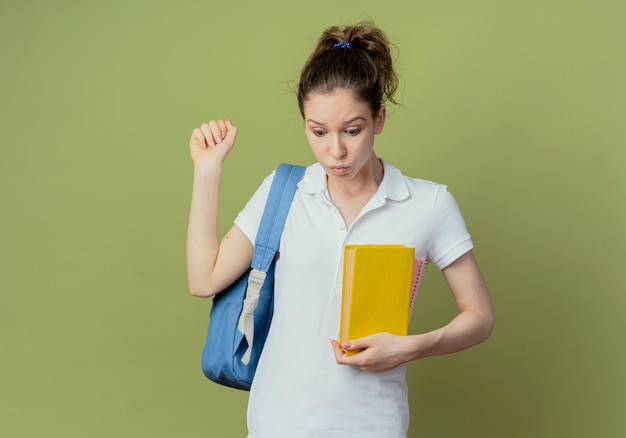 Impressed young pretty female student wearing back bag looking down holding book and clenching fist isolated on green background with copy space